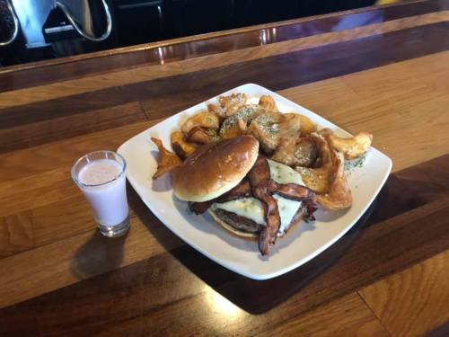 A plate with a burger topped with bacon and cheese, served with seasoned potato wedges and a small glass of pink drink.