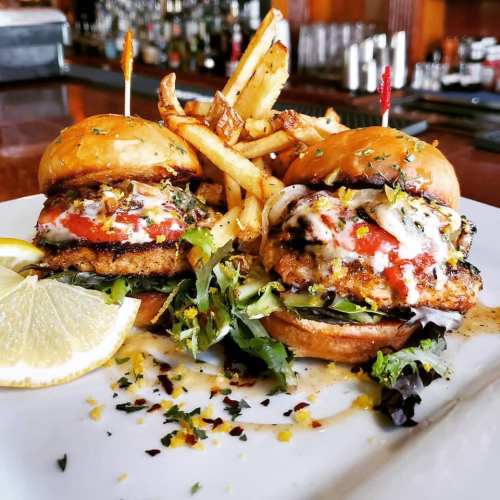 Two chicken sliders topped with sauce and cheese, served with fries and a lemon wedge on a white plate.