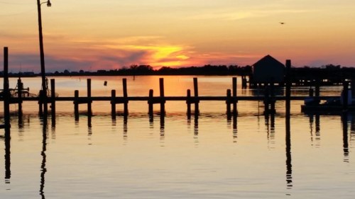 A serene sunset over calm waters, reflecting vibrant orange and purple hues, with a dock and silhouettes of boats.