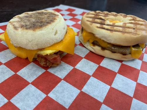 Two breakfast sandwiches on a red and white checkered paper: one with bacon and cheese, the other with sausage and cheese.