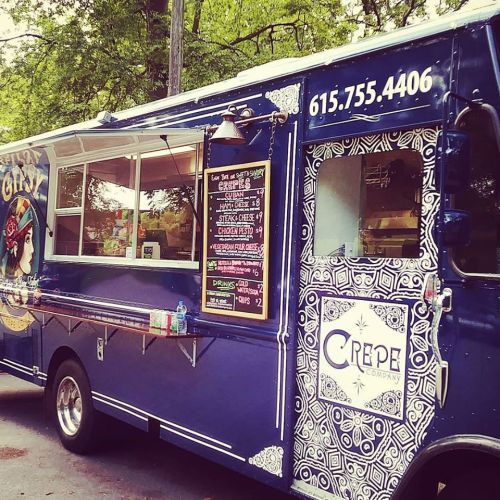 A blue food truck with a crepe menu and contact number displayed on the side, parked in a green area.