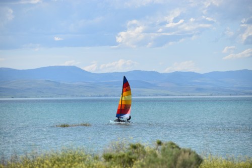 A colorful sailboat glides across a calm lake with mountains in the background under a partly cloudy sky.