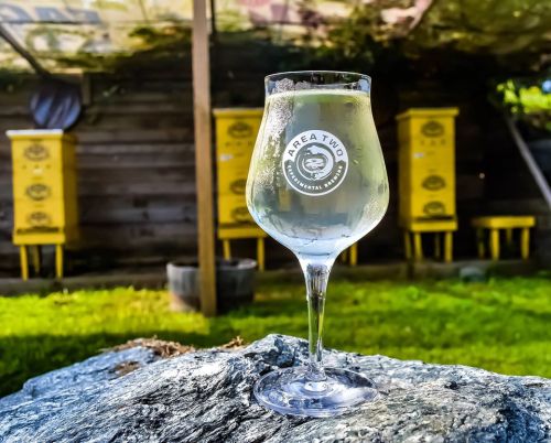 A glass of clear beverage sits on a rock, with yellow beehives and greenery in the background.