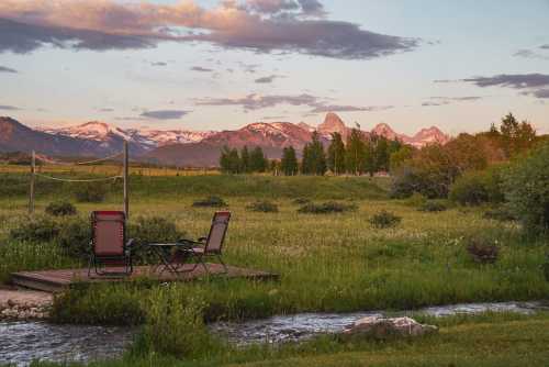 A serene landscape featuring two chairs on a wooden deck by a stream, with mountains and a colorful sunset in the background.