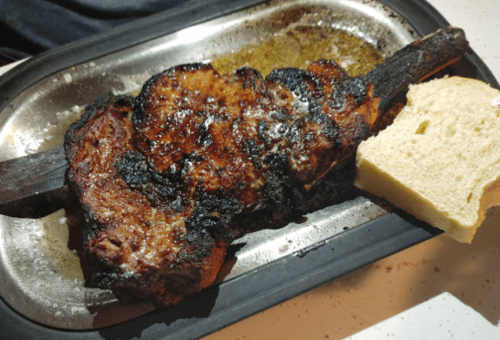 A grilled piece of meat on a metal platter, accompanied by a slice of bread.