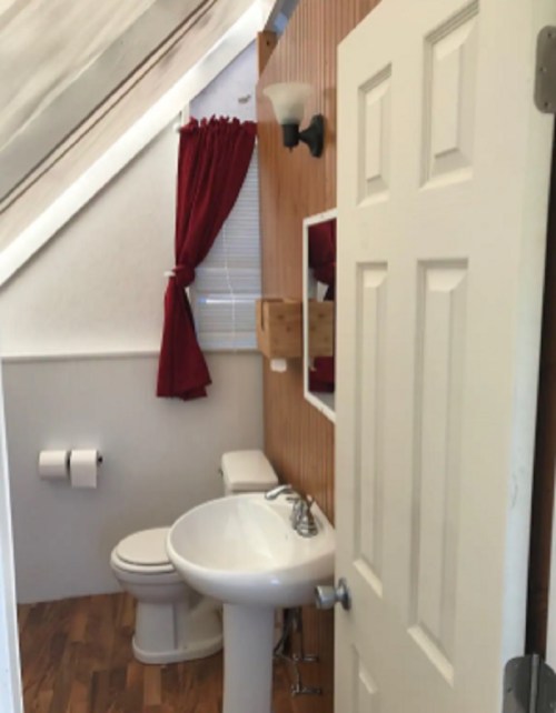A small bathroom with a sink, toilet, and wooden accents, featuring red curtains and a door partially open.