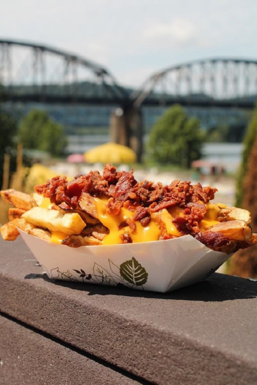 A close-up of loaded fries topped with cheese and bacon, with a bridge and trees in the background.