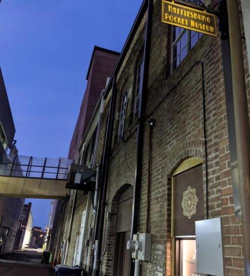 A brick building at dusk with a sign for the Hattiesburg Pocket Museum and a walkway above.