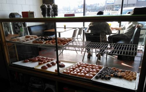 A display case filled with various donuts, with customers seated in the background of a bright cafe.