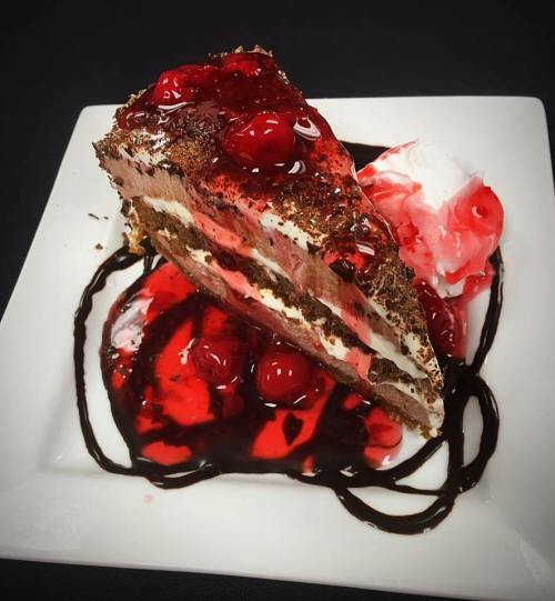 A slice of chocolate cake layered with cream and cherries, drizzled with chocolate sauce and served with whipped cream.