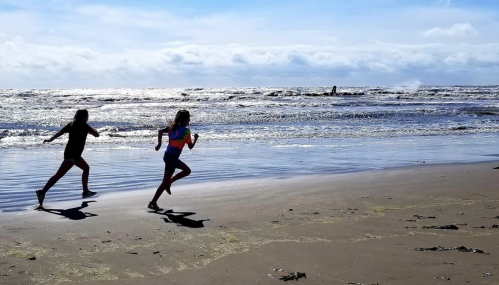 Two children run along a sandy beach, with waves crashing in the background under a bright blue sky.