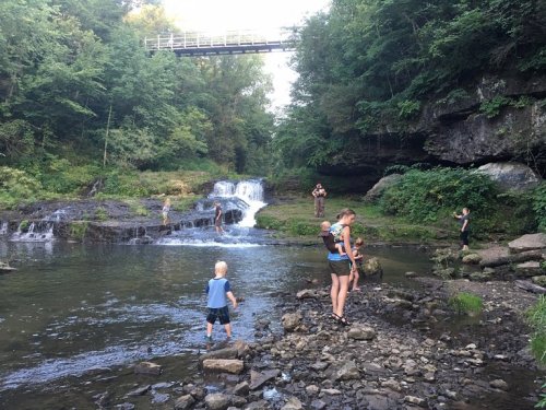 A group of people enjoying a river with a waterfall, surrounded by lush greenery and a bridge overhead.