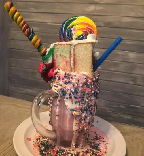 A colorful milkshake topped with whipped cream, a slice of cake, a lollipop, and a candy stick, overflowing with sprinkles.