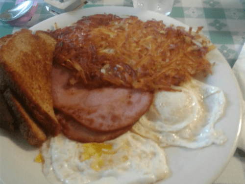 A plate of breakfast featuring hash browns, ham slices, fried eggs, and toast.