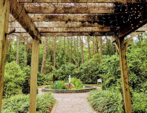 A wooden pergola with lights leads to a serene garden path surrounded by greenery and a white cross in the distance.