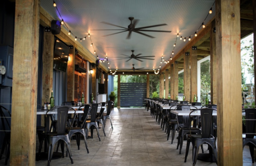 A spacious outdoor dining area with wooden beams, ceiling fans, and string lights, featuring empty metal chairs and tables.