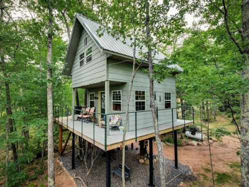 A modern treehouse with a green exterior, elevated on stilts, surrounded by lush trees and a small patio area.