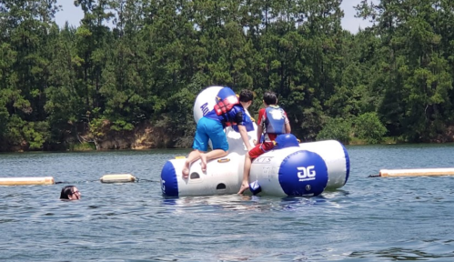 Two kids climb onto a floating inflatable structure in a lake while another swims nearby. Lush trees line the shore.