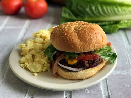 A juicy burger with lettuce, tomato, and bacon, served with a side of scrambled eggs on a plate. Fresh produce in the background.