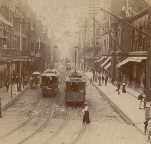 Historic street scene with trams, pedestrians, and buildings, capturing a bustling urban atmosphere from the past.