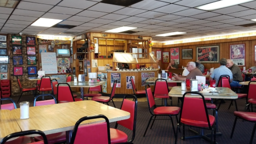A cozy diner interior with red chairs, wooden tables, and vintage posters on the walls. Two patrons are seated.