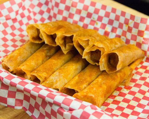 A basket lined with red and white checkered paper filled with golden-brown taquitos.