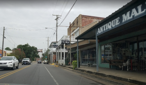 A street view featuring shops, including an antique mall, with cars parked along the road and a cloudy sky above.