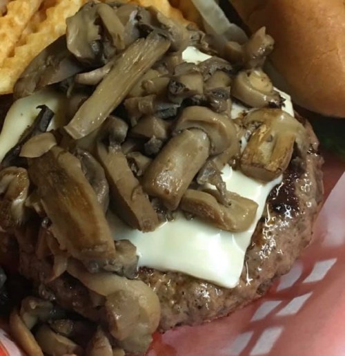 A close-up of a burger topped with sautéed mushrooms and melted cheese, served with waffle fries and a bun.