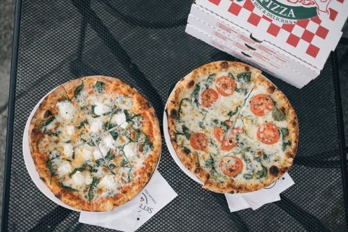 Two pizzas on a table: one with spinach and cheese, the other topped with tomatoes and herbs, next to a pizza box.