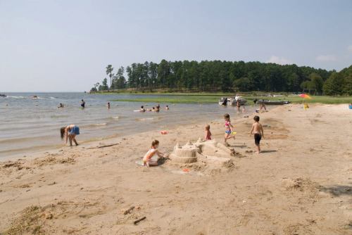 Children playing on a sandy beach, building a sandcastle, with people swimming and boats in the background.