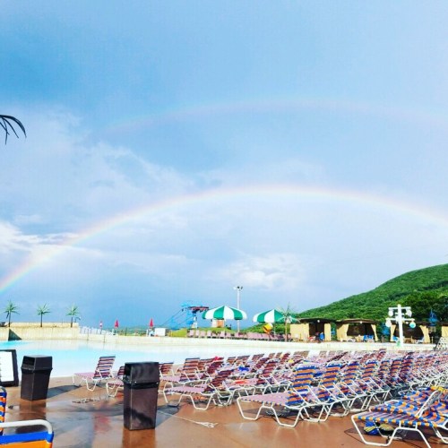 A pool area with colorful lounge chairs, under a blue sky featuring two rainbows and a green hillside in the background.