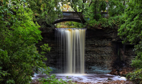 A serene waterfall cascades over rocky cliffs, surrounded by lush green trees and a stone bridge above.