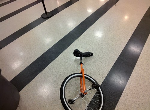 An orange unicycle lies on a striped floor in a public space.