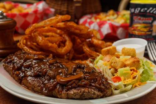 A plate of steak topped with mushroom gravy, served with onion rings, salad, and croutons.