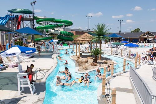 A vibrant water park scene with a lazy river, water slides, palm trees, and people enjoying the sun and water activities.