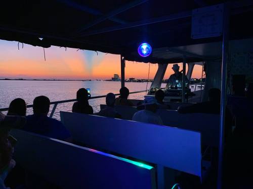 A group of people on a boat watching a sunset over the water, with city lights in the background.