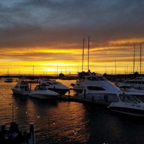 Sunset over a marina with yachts docked, reflecting golden hues on the water and a cloudy sky.
