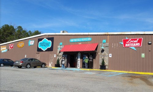 Exterior of a shop with a red awning, signs for local artisans, and a parking lot under a clear blue sky.