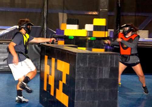 Two players in protective gear engage in a laser tag game, taking cover behind a colorful block structure.