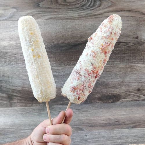 A hand holding two corn on the cob popsicles, one coated in white and the other in red seasoning, against a wooden background.