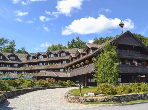 A rustic lodge with multiple levels, surrounded by greenery and blue skies, featuring flower boxes and a stone pathway.