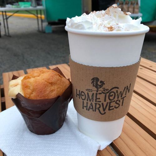 A coffee cup topped with whipped cream and a muffin on a wooden table, with a logo reading "Hometown Harvest."