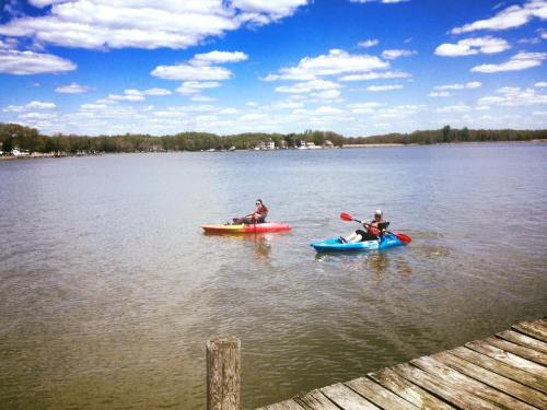 Two people kayaking on a calm lake under a blue sky with fluffy clouds, with a wooden dock in the foreground.