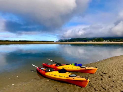 Two kayaks, one orange and one yellow, rest on a sandy shore beside a calm, reflective water under a cloudy sky.