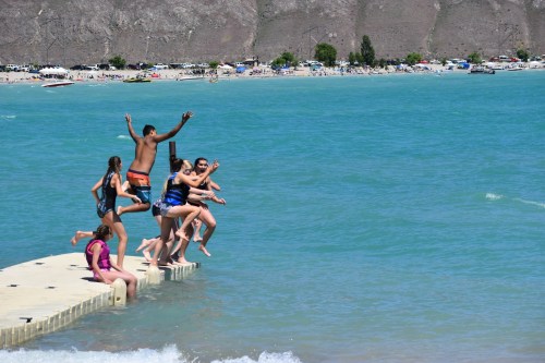 A group of children and teens jumping off a dock into a turquoise lake on a sunny day.