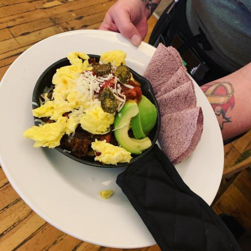A person holds a plate with a skillet dish featuring scrambled eggs, cheese, salsa, jalapeños, avocado, and tortillas.