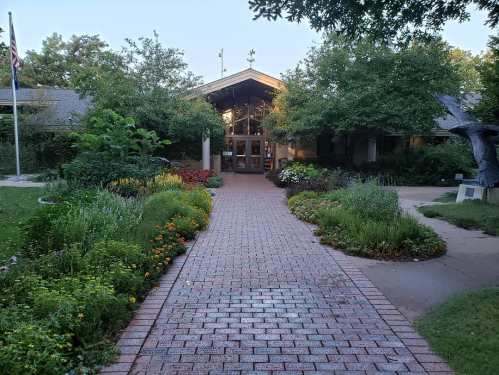 A brick pathway leads to a welcoming building surrounded by lush greenery and colorful flower beds.