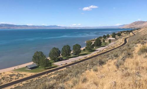 A scenic view of a lake with trees and a winding road along the shore, surrounded by hills and blue skies.