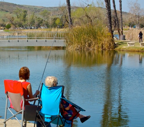 Two people sit in folding chairs by a calm lake, fishing with a bridge and lush greenery in the background.