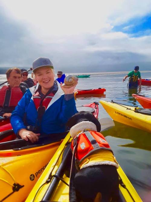 A smiling woman holds a shell while kayaking with others; a dog in a life jacket sits beside her in a colorful kayak.